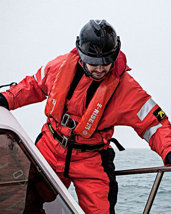 Crew member in a red flotation suit and lifejacket holding a boat rail